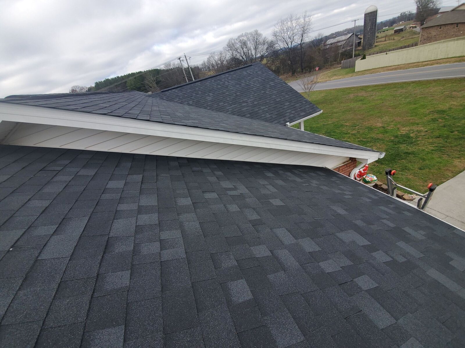 Finished dark architectural shingle roof seen from roof level on an overcast winter day, with bare trees and farmland behind