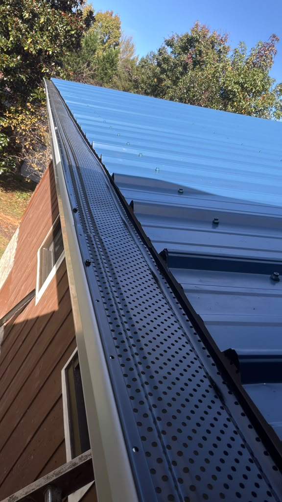 New black gutter guard installed along the edge of a blue metal roof, with fall trees behind it